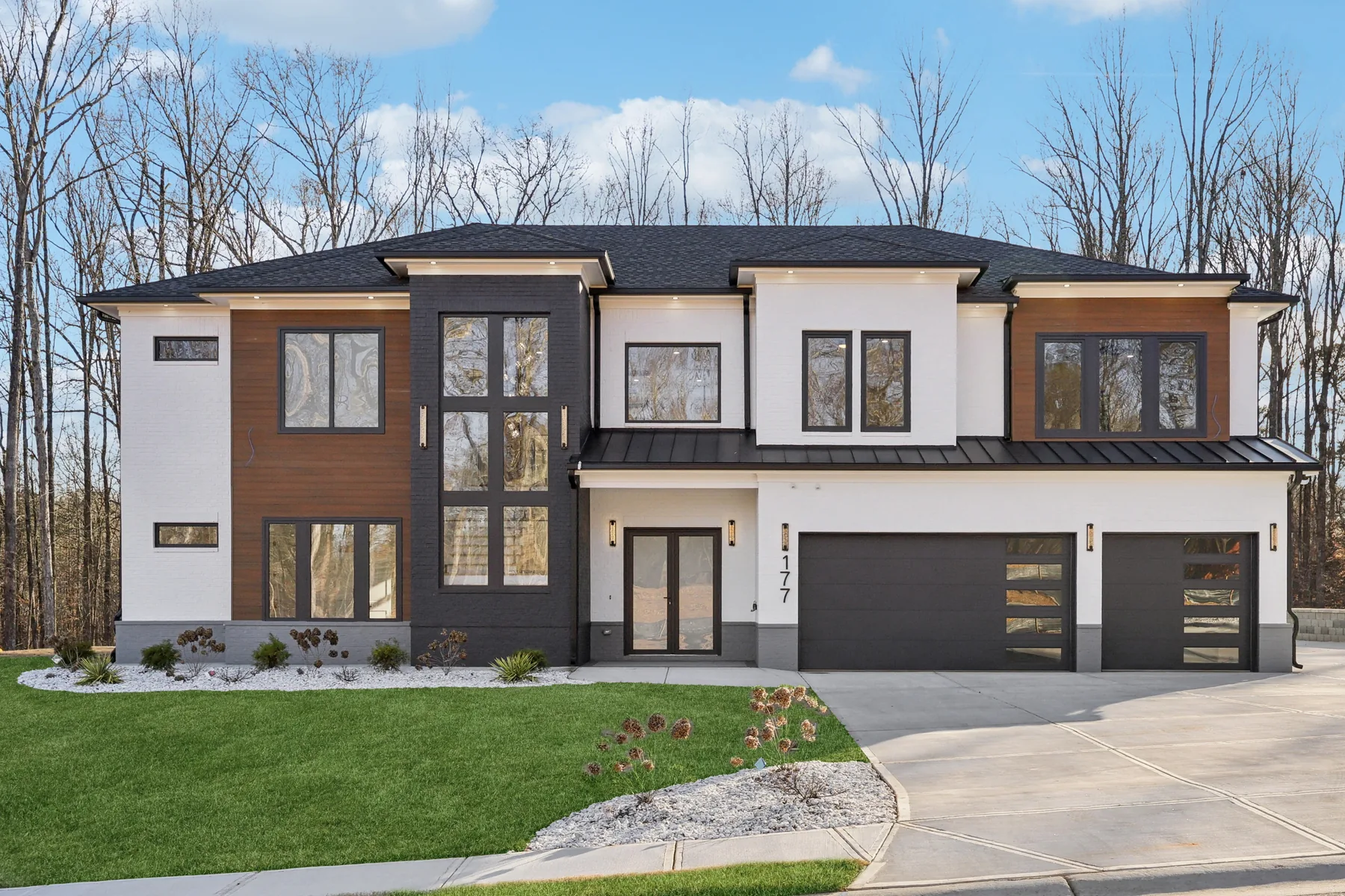 Front elevation of the Deer Canyon home: two-story modern facade in white stucco with dark wood accent panels, black window frames, a flat parapet roofline, and a wide concrete driveway
