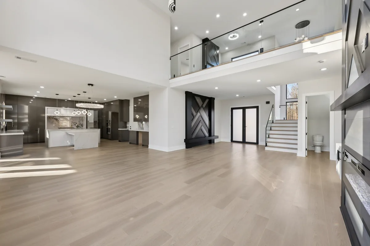 Double-height great room with wide-plank flooring, a full wall of black-framed windows, and an open walkway across the second floor