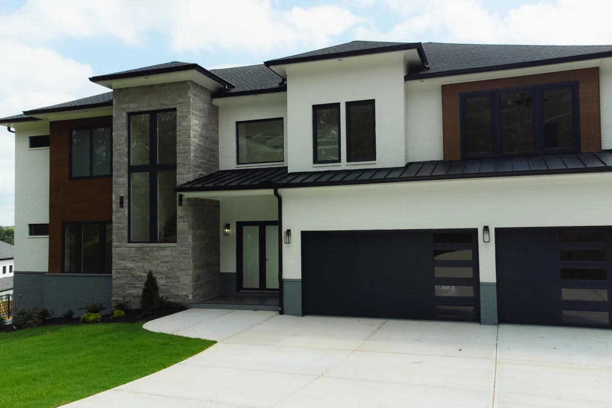 Front elevation of the 670 Moon Place Rd home: white stucco with stone veneer and vertical wood accent panels under a wide hipped roof with deep eaves, black window frames, and a three-car garage