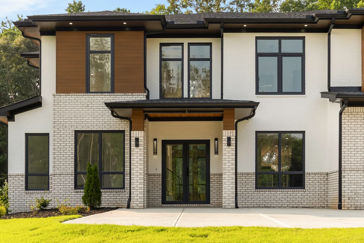 Front elevation in daylight: painted brick with wood accent panels, black window frames, and a three-car garage set into a sloped lot