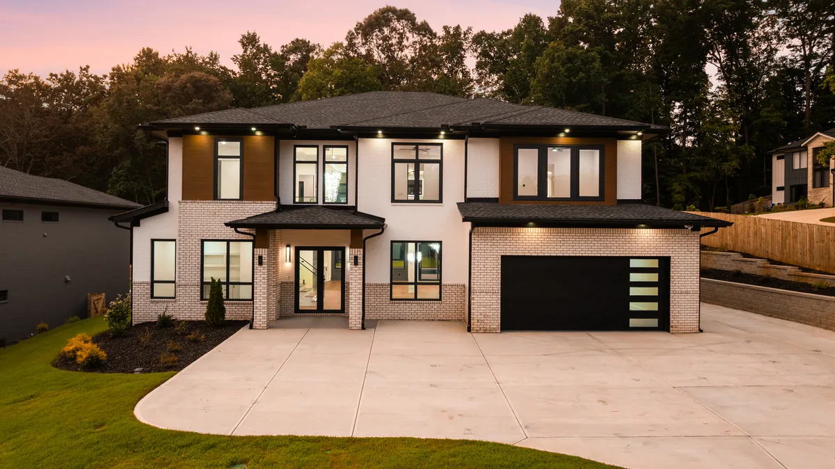 Front elevation photographed at dusk under a pink sky, showing the painted brick and stone facade, dark garage doors, and a wood-clad entry tower