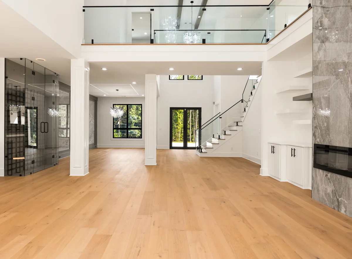 Double-height great room with white oak plank flooring, a floor-to-ceiling marble fireplace surround, a glass-enclosed wine room, and an open second-floor walkway above