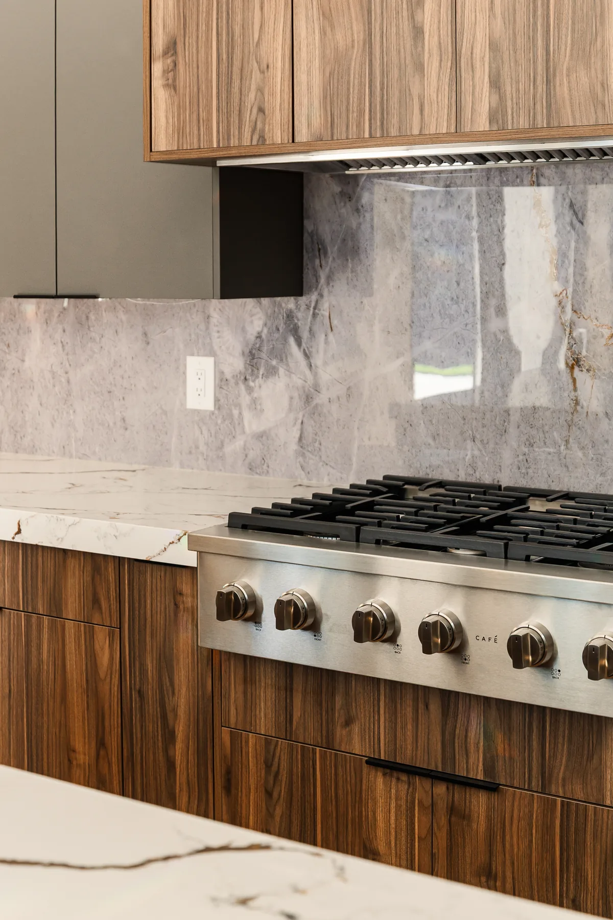 Cooking wall with a professional gas range, wood-panelled cabinetry, and a bookmatched marble-look slab backsplash running to the hood