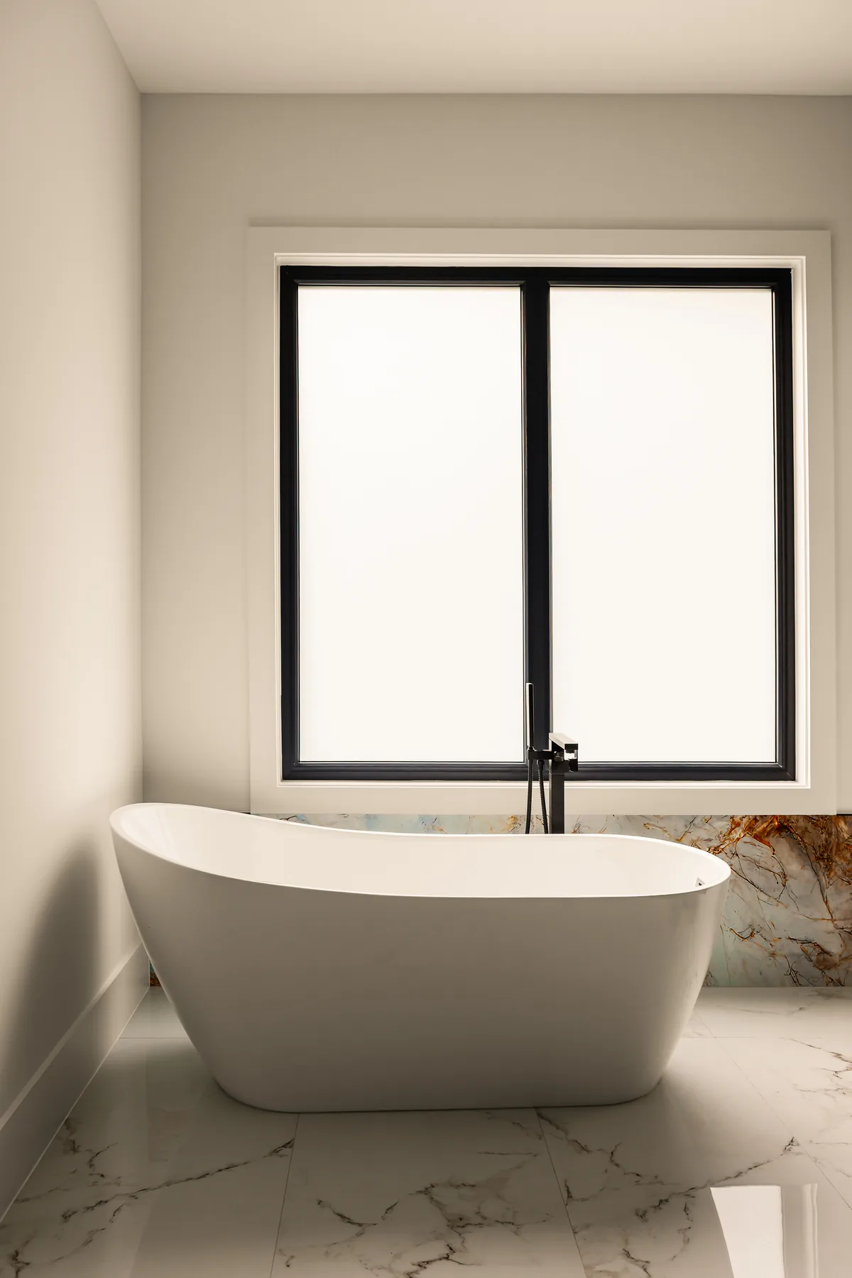 Freestanding white soaking tub set against a plain wall beneath a tall black-framed window