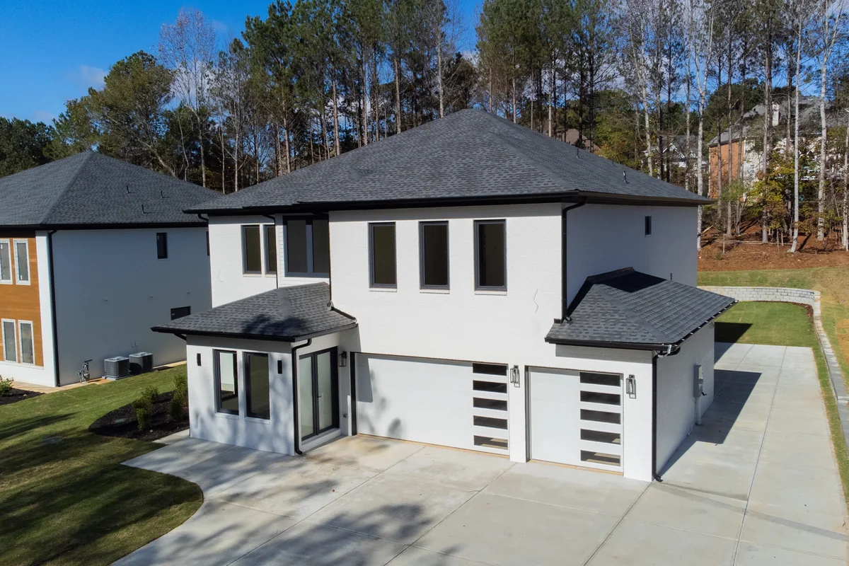 The front of the house seen at an angle from the driveway, showing the garage wing, the entry porch, and the wooded tree line behind