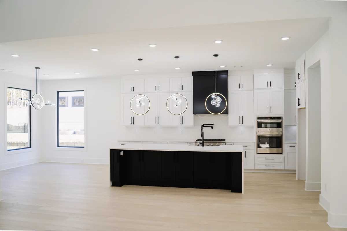 Wide view of the kitchen from the living area: a dark island against white perimeter cabinetry, pale oak flooring, and a black hood