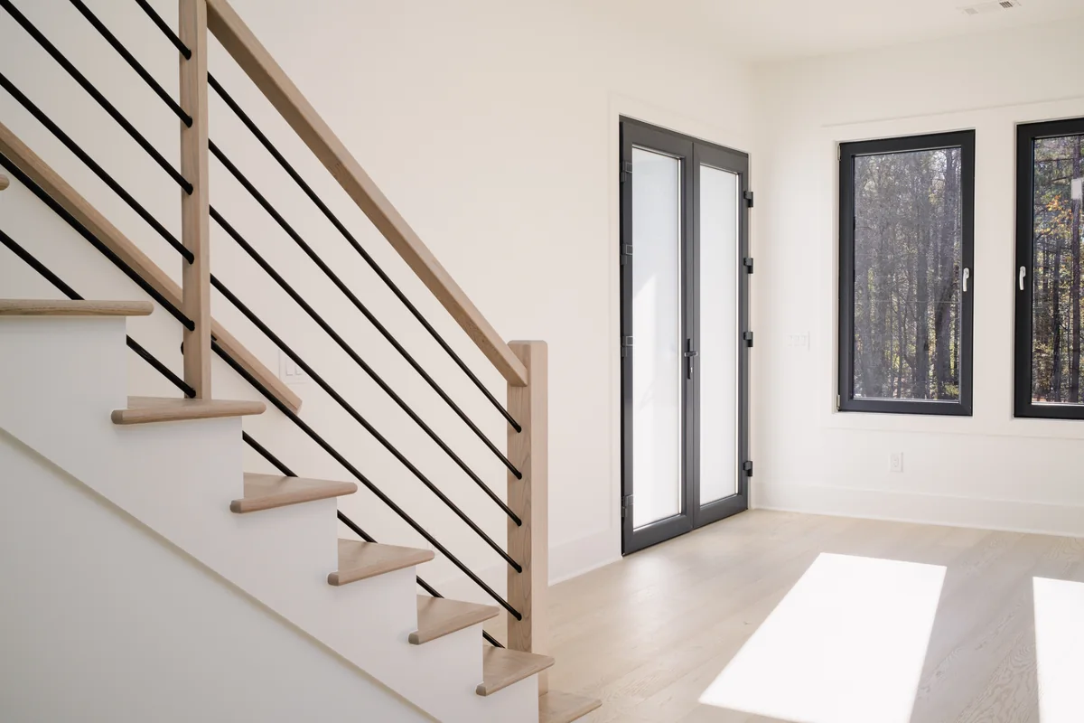 Detail of the stair with its black horizontal metal rail and oak handrail against a white wall, with a black-framed entry door beyond