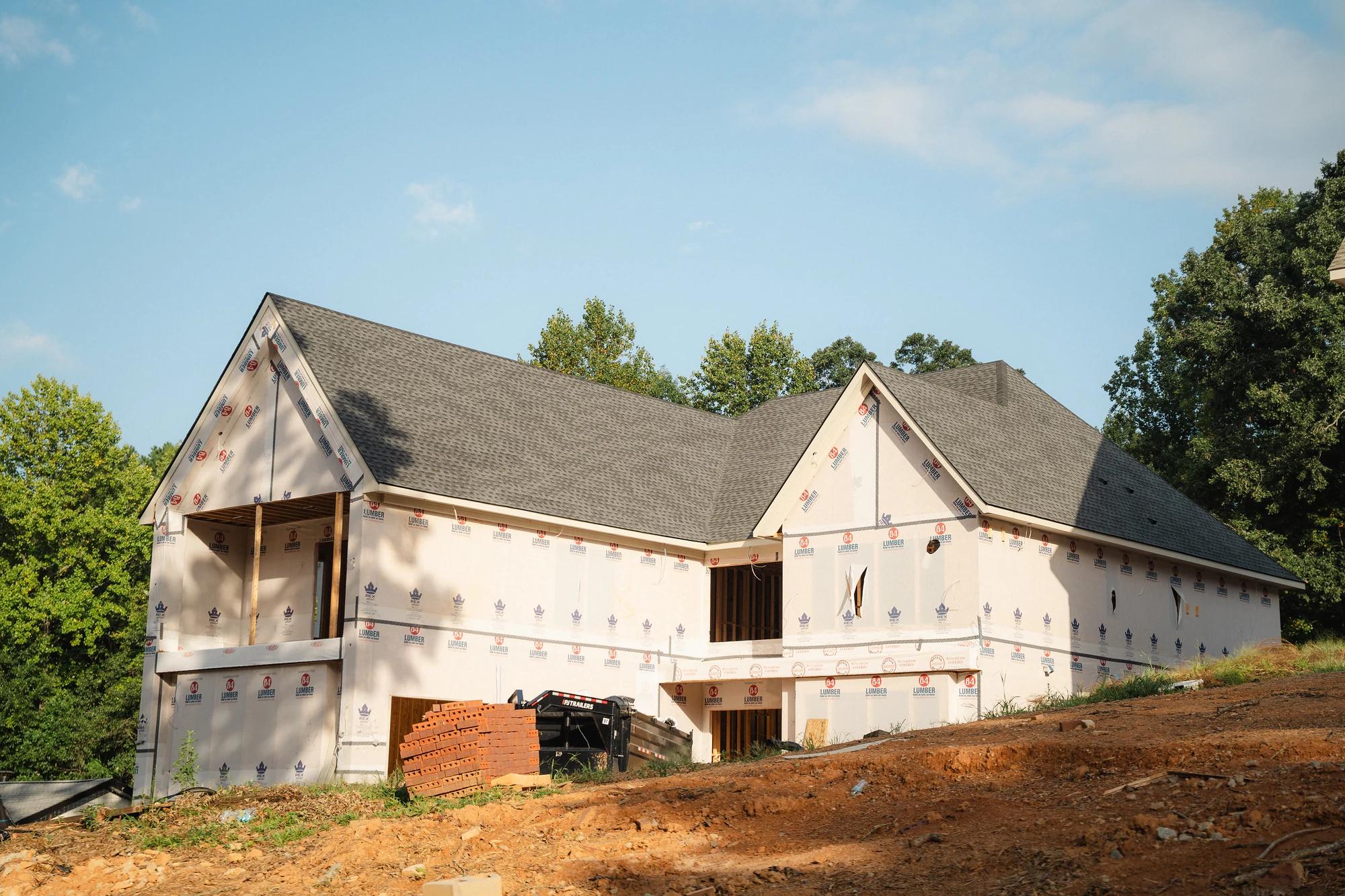 Wide exterior view of a roofed house wrapped in weather barrier on a graded dirt site.