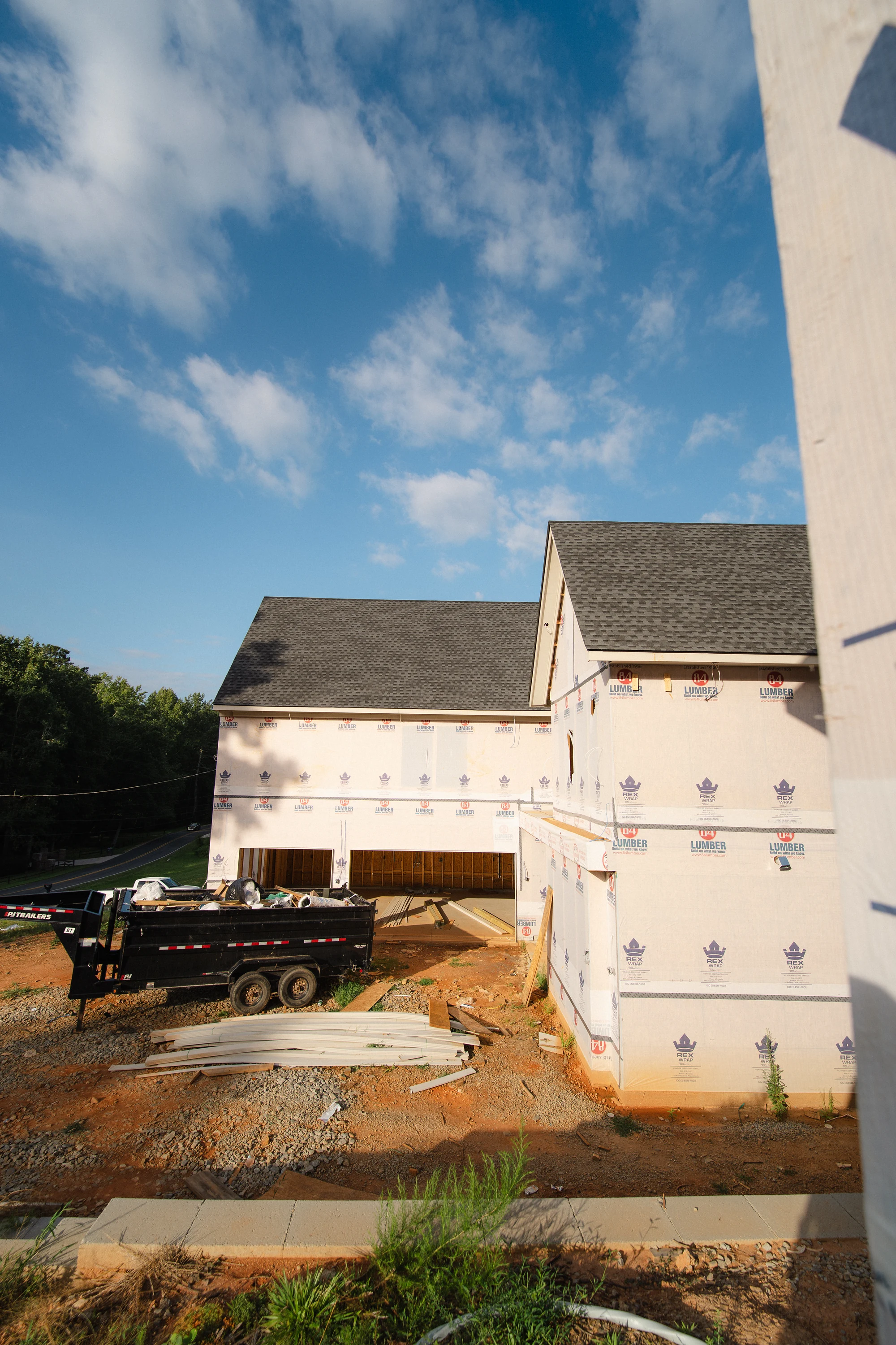 Side exterior with shingled gables, weather-wrapped walls, open lower bays, and a site trailer.