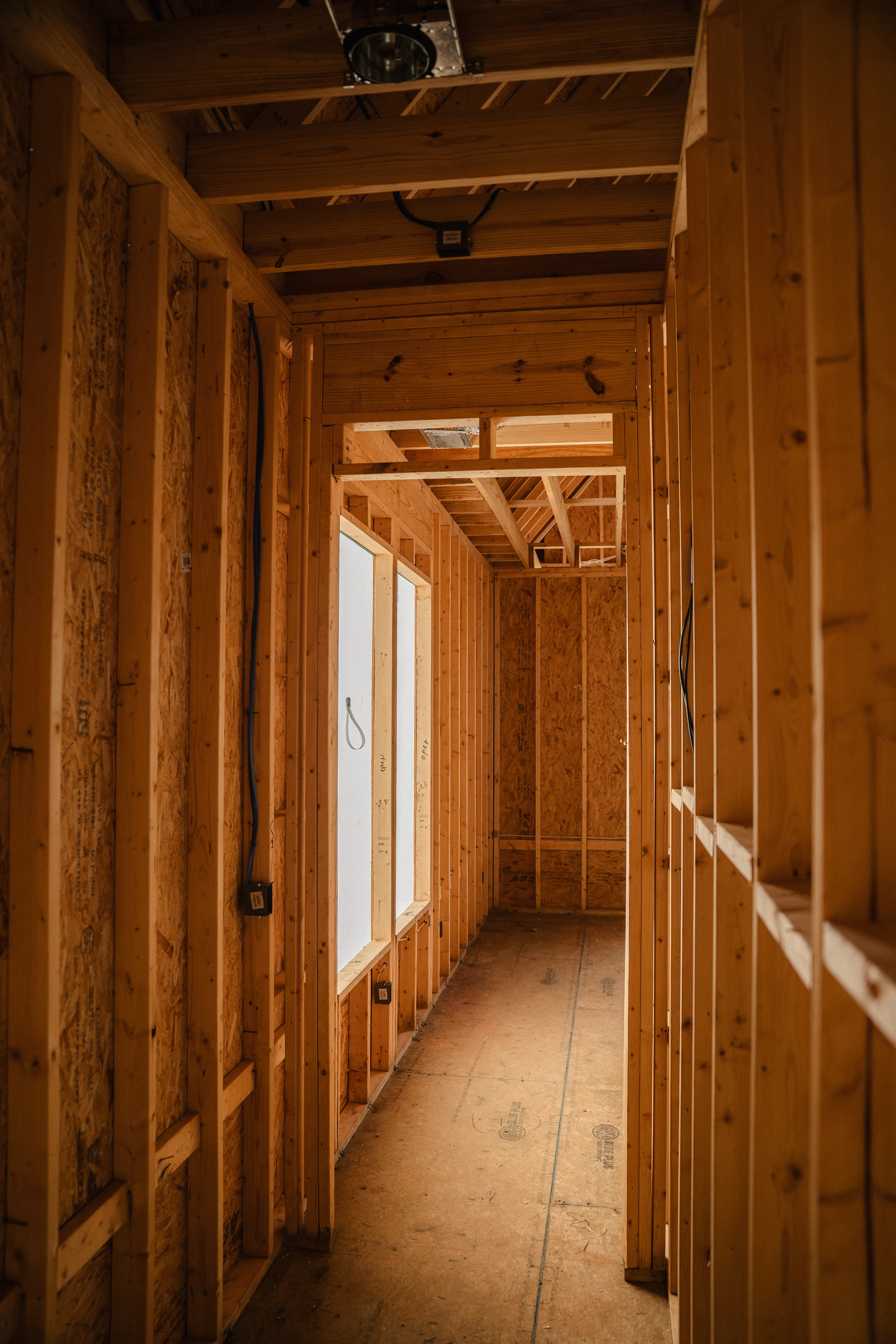 Narrow framed corridor with exposed studs, ceiling joists, wiring, and a bright opening ahead.