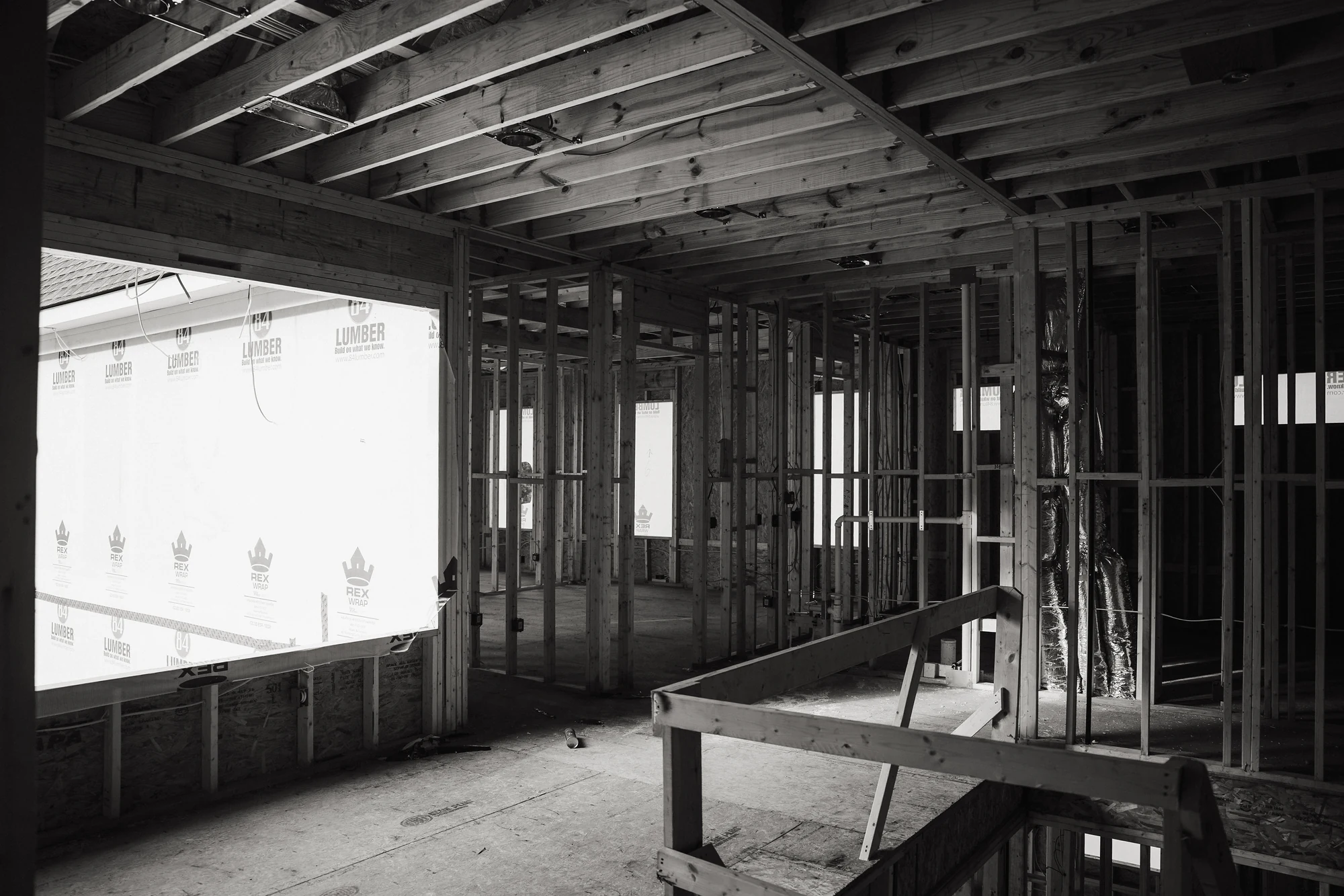 Black-and-white interior showing exposed joists, studs, a large exterior opening, and temporary rails.