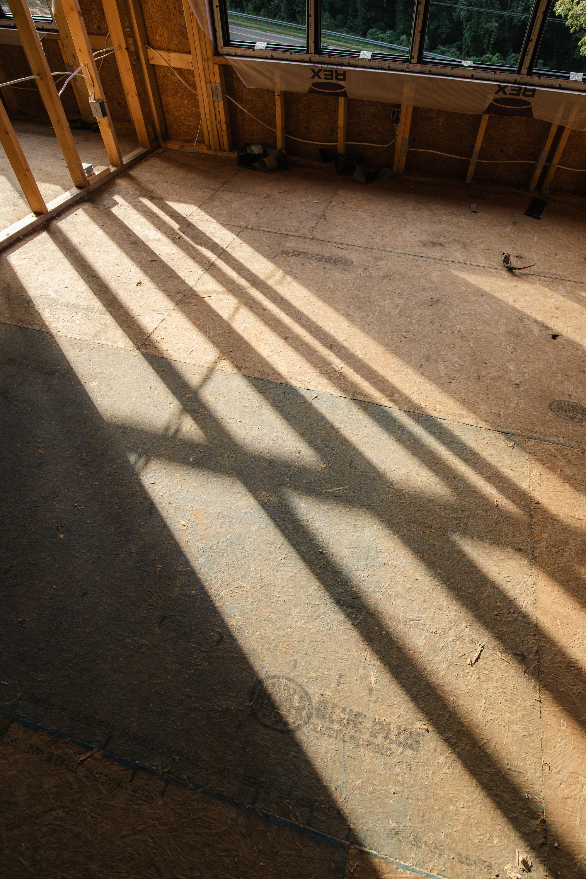 Long shadows from wall framing cross an unfinished wood subfloor below installed windows.