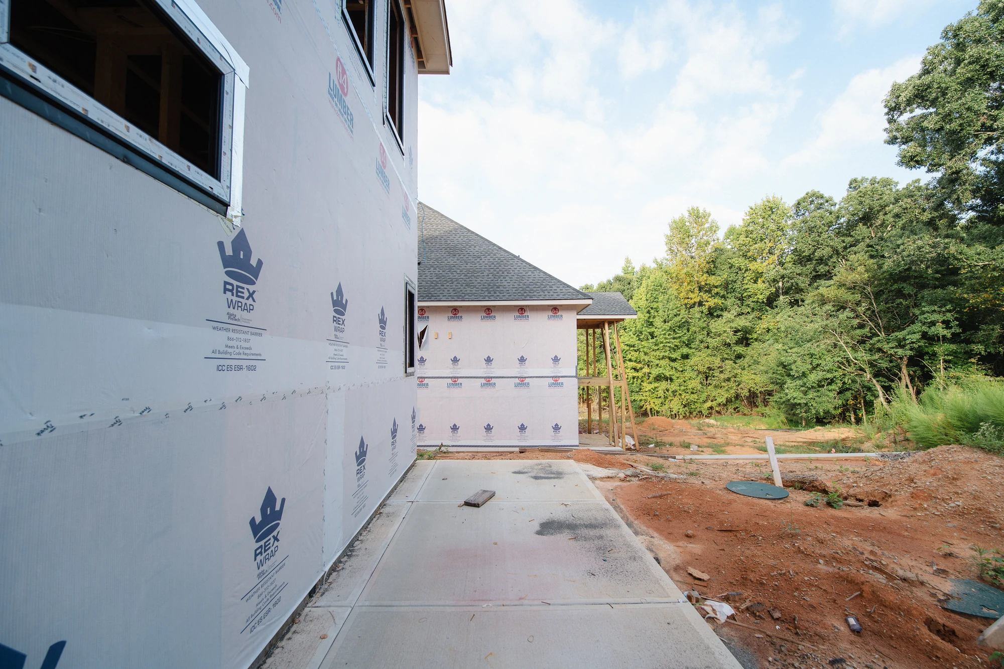 Concrete side walkway between weather-wrapped walls and a graded dirt area with utility pipes.