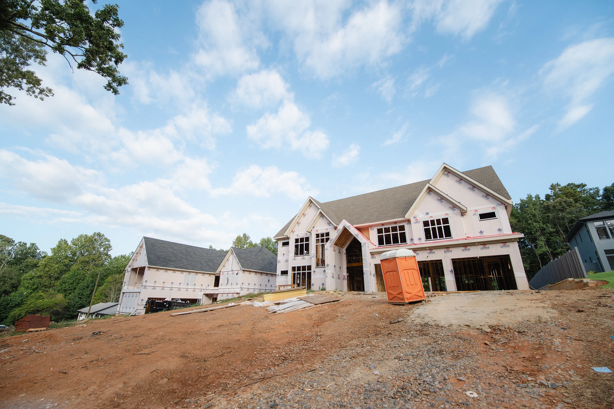 Wide street-side view of a large roofed house on a graded dirt lot with wrapped exterior walls.