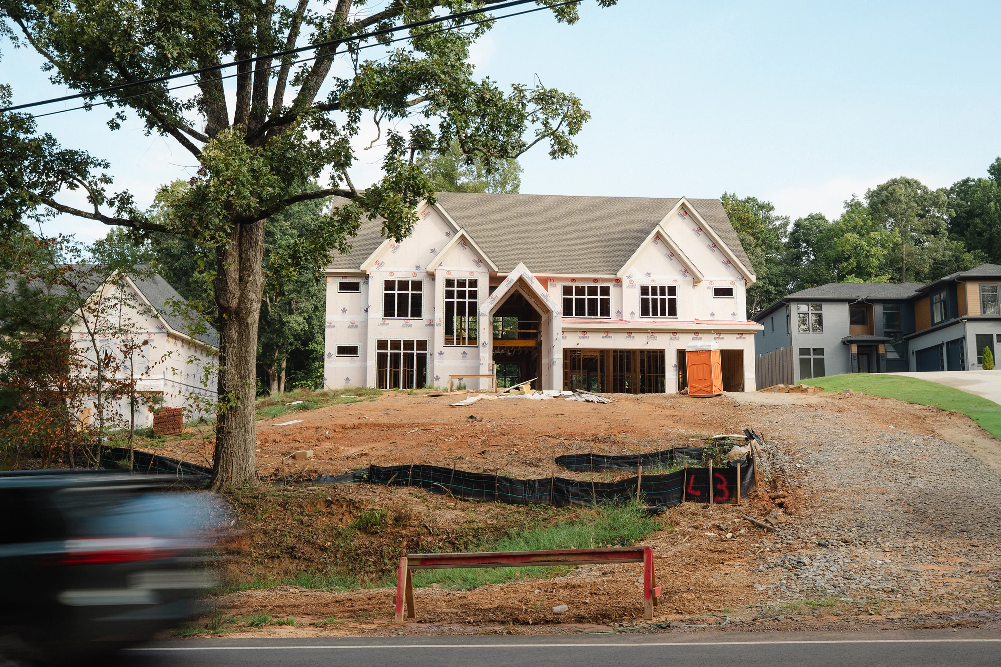 Street view through mature trees of a roofed house under construction on a sloped site.