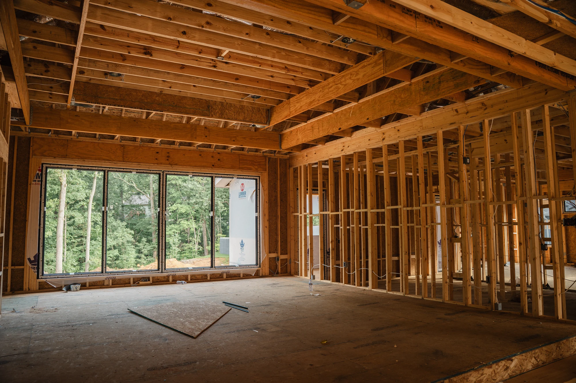 Broad framed interior with exposed joists, wall studs, subfloor, and a large window opening.