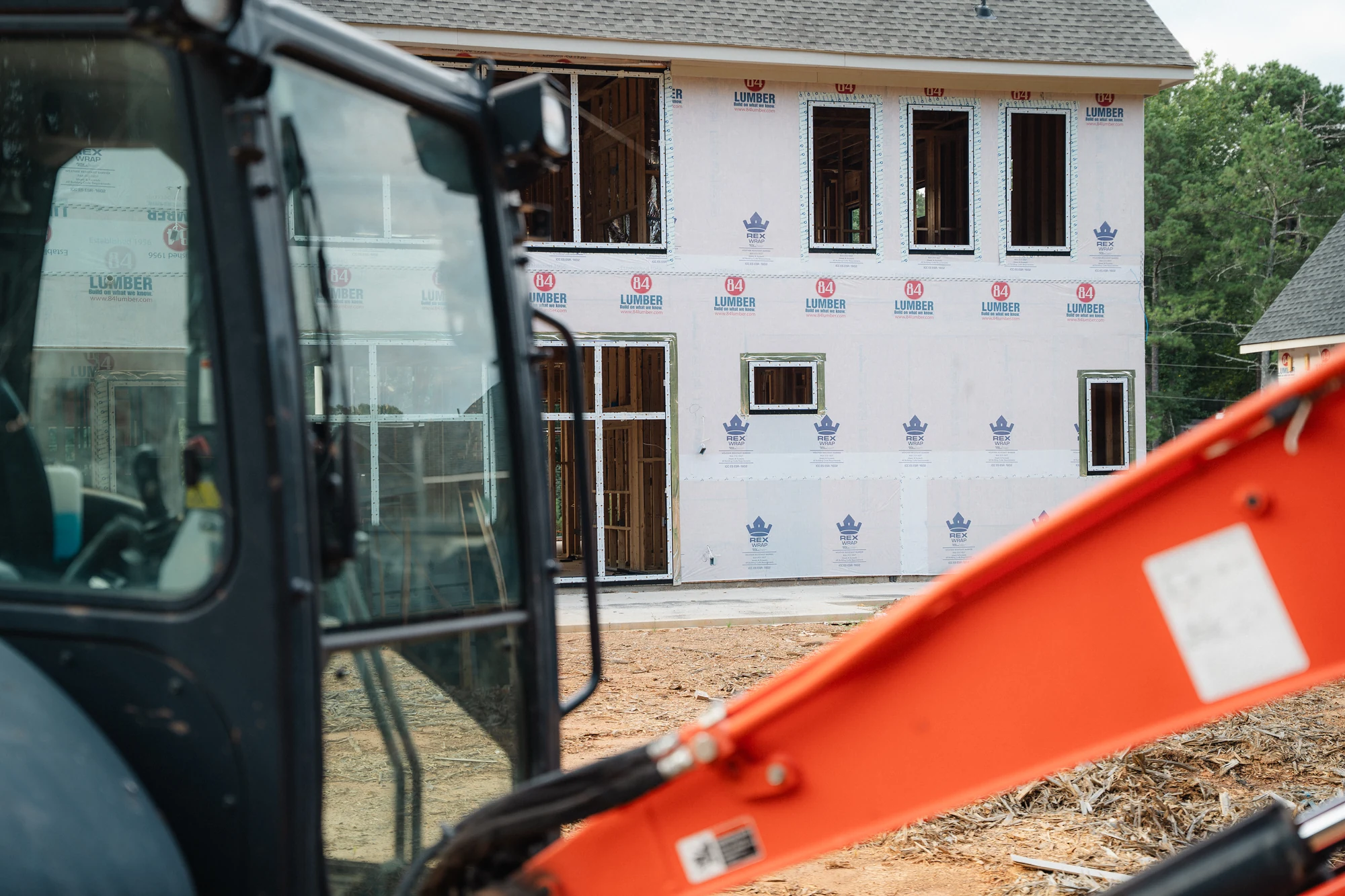 Construction equipment in front of a weather-wrapped two-story exterior with window openings.